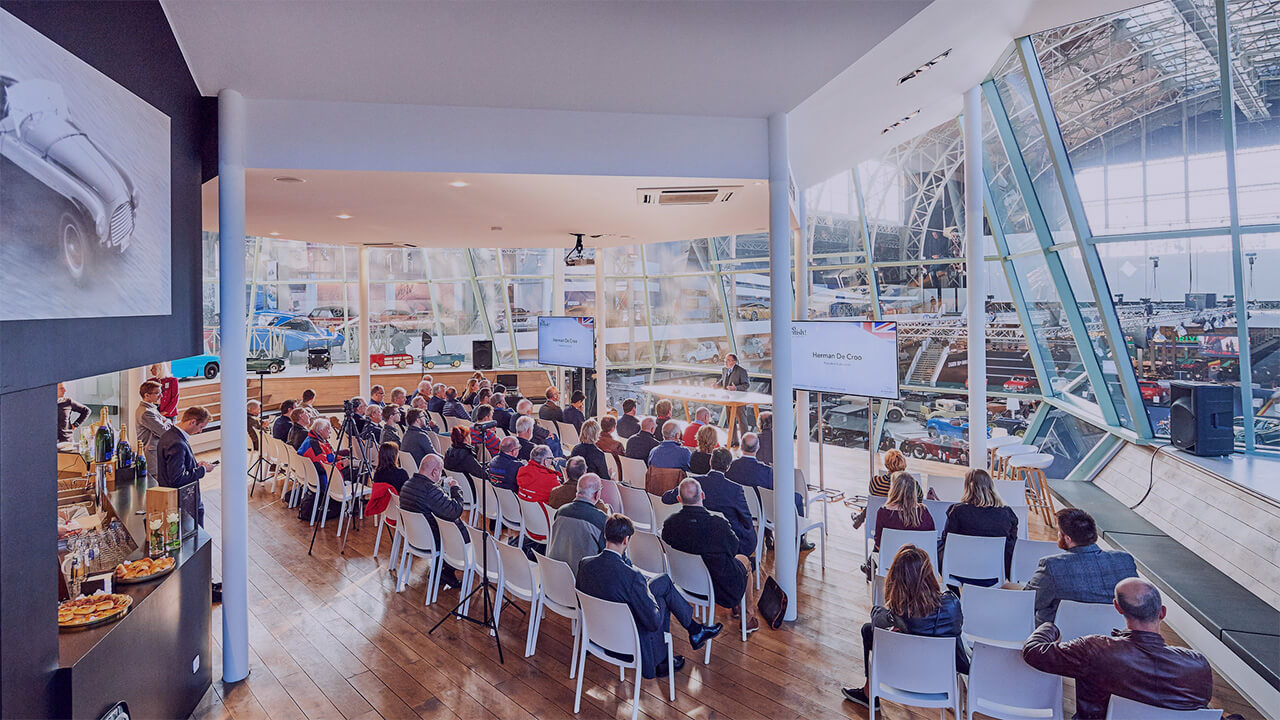 Conférence dans le Classic Lounge d'Autoworld Bruxelles, avec une audience attentive et une vue panoramique sur l'exposition de voitures anciennes en arrière-plan.