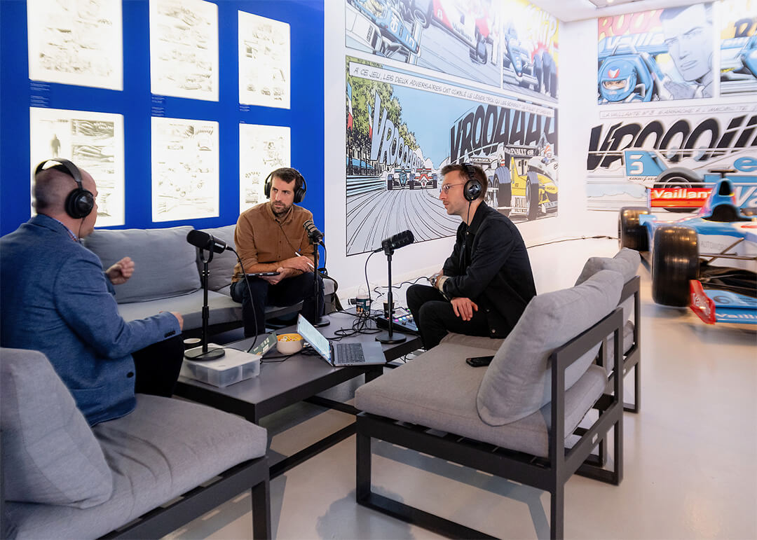 Trois hommes enregistrent un podcast dans un studio décoré de planches de bande dessinée et d’une voiture de course exposée.