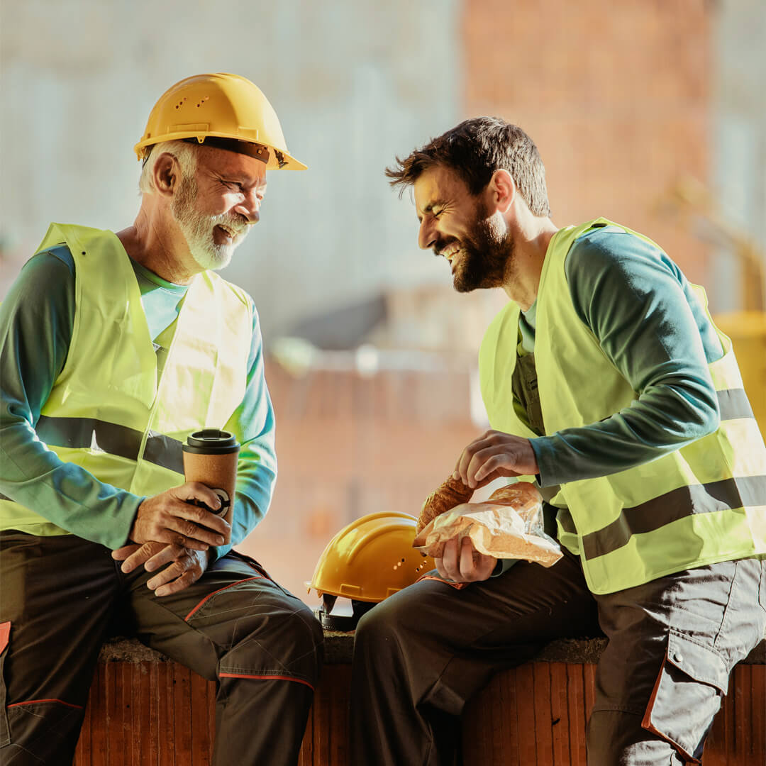 Deux ouvriers en gilet fluorescent partagent un moment convivial lors d’une pause sur un chantier.